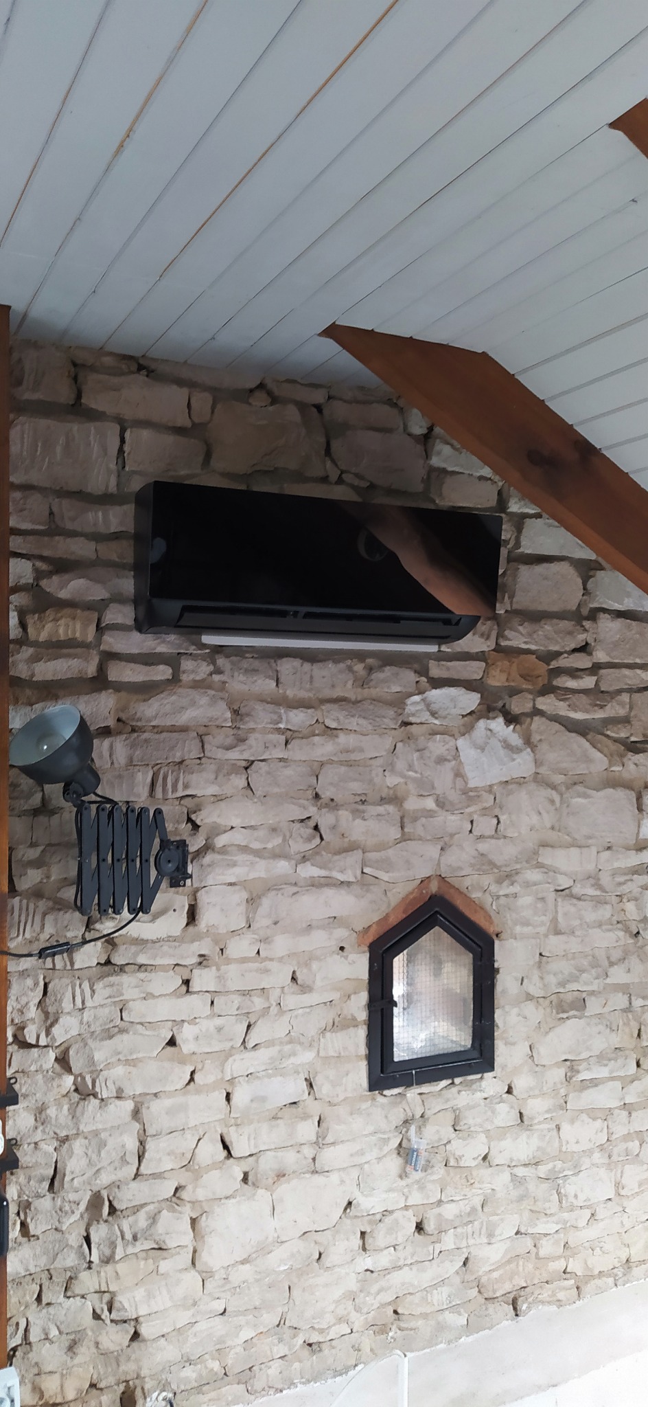 Czarny klimatyzator ścienny zamontowany na ścianie z jasnego kamienia, widoczna lampa na teleskopowym ramieniu i małe okienko z kratką na ścianie, drewniany element konstrukcyjny pod białym...