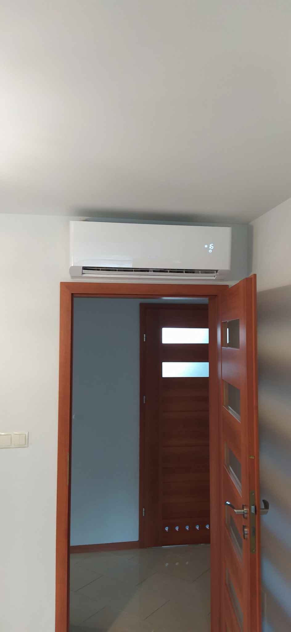 Biały klimatyzator ścienny zamontowany nad otwartymi, drewnianymi drzwiami wewnętrznymi z przeszkleniami, widoczny wyświetlacz temperatury.