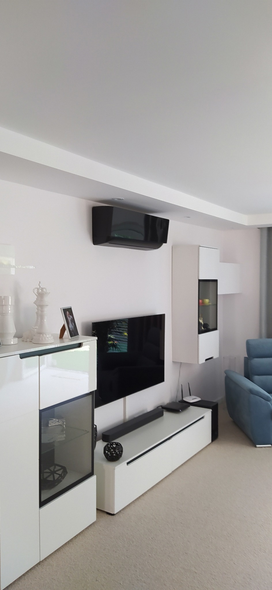 Czarny klimatyzator ścienny w nowoczesnym salonie z białymi meblami i telewizorem.