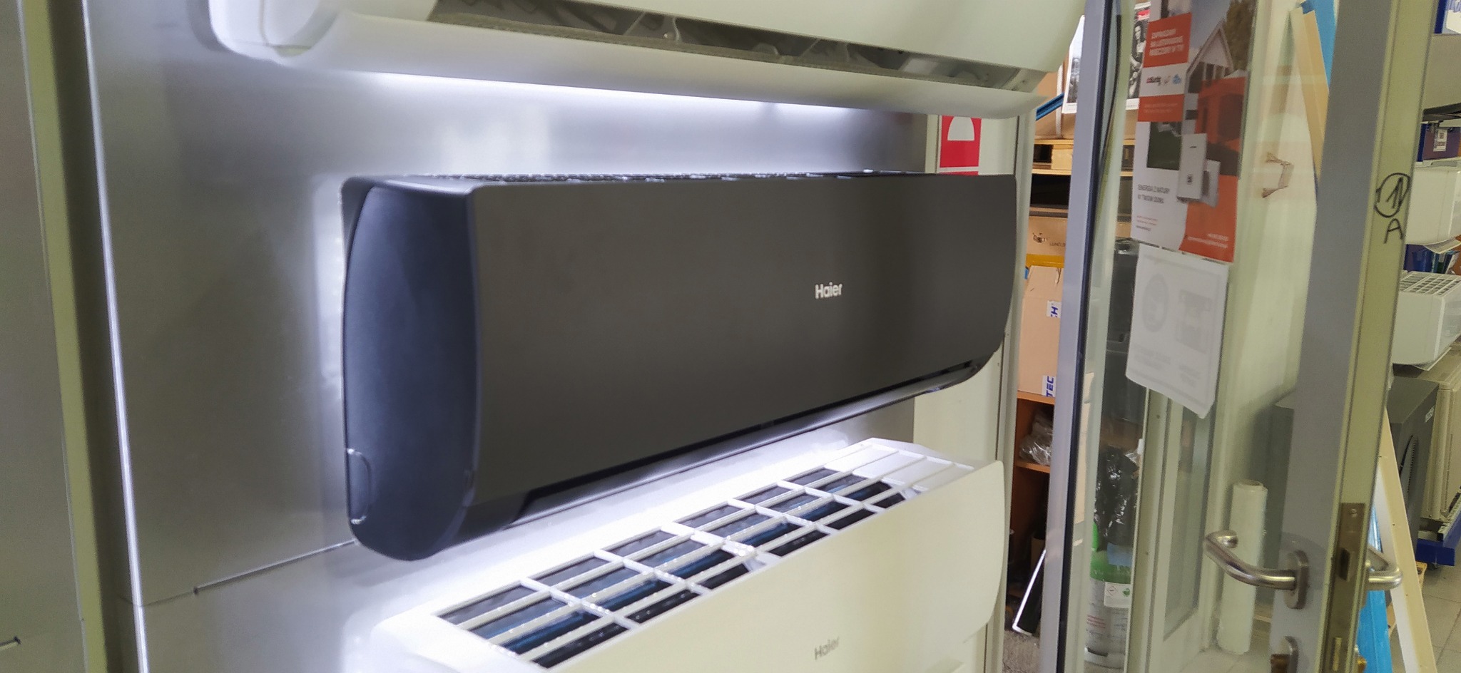 Wystawa klimatyzatorów Haier w sklepie, w tym model w kolorze grafitowym i białe jednostki, widoczne przez otwarte drzwi sklepu