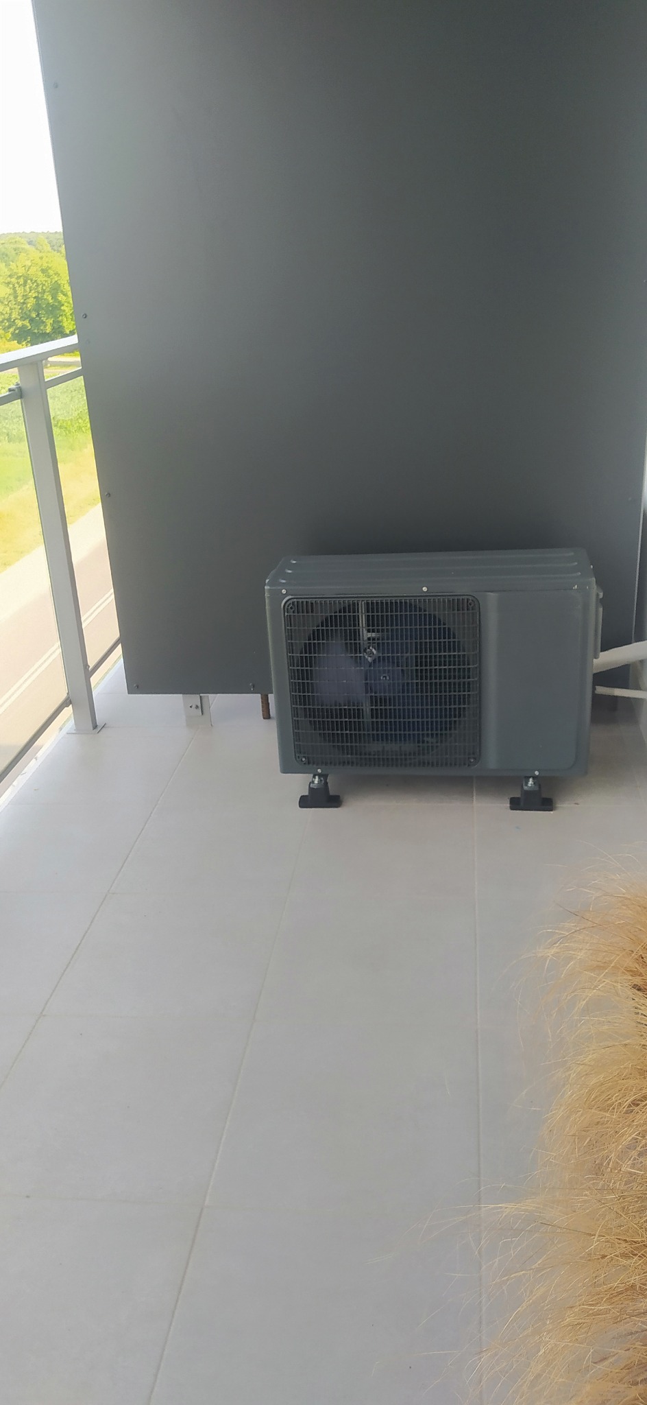 Szara jednostka zewnętrzna klimatyzacji na balkonie z widokiem na zieleń przez balustradę.