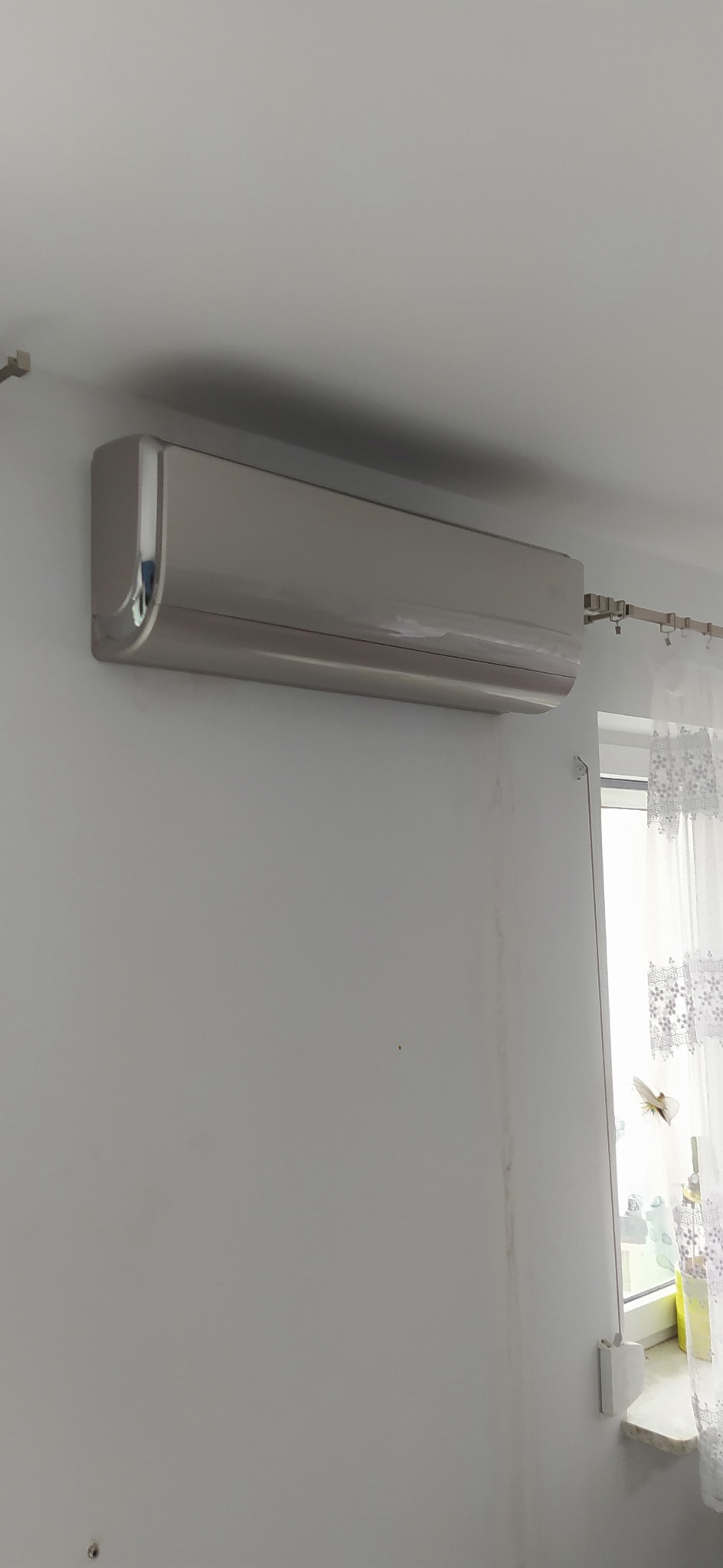 Nowoczesny, srebrny klimatyzator ścienny w jasnym pomieszczeniu z widocznymi śladami po montażu na ścianie i firanką w oknie.