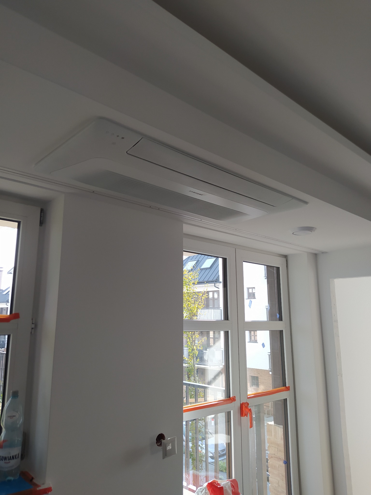 Sufitowa jednostka klimatyzacyjna Samsung zamontowana w jasnym pomieszczeniu z oknami, zabezpieczonymi pomarańczową folią malarską.