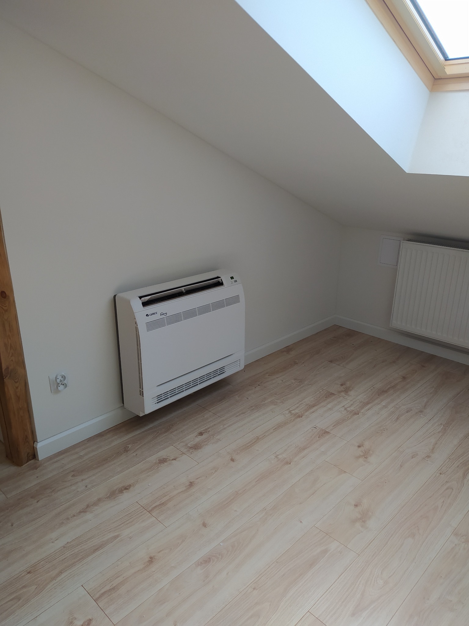 Biały klimatyzator podłogowy Gree zainstalowany w jasnym pokoju na poddaszu z oknem dachowym i panelami podłogowymi.
