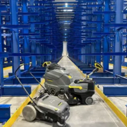 Trzy urządzenia czyszczące Kärcher, w tym zamiatarka z szczotką, automat szorujący i mała szorowarka, stoją na betonowej posadzce w magazynie z wysokimi, niebieskimi regałami.