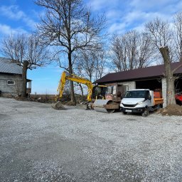 IZDEBSKI - Żółta koparka, walec i biała ciężarówka Iveco na utwardzonym żwirowym placu budowy, w tle budynki gospodarcze i drzewa bez liści, pochmurne niebo.