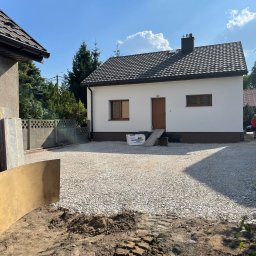 IZDEBSKI - Podwórko z nowo wysypanym żwirem przed białym domem z brązowymi oknami i drzwiami, widoczne ślady opon i deska oparta o ścianę budynku gospodarczego.