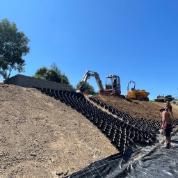 IZDEBSKI - Skarpa wzmocniona geokratą i zabezpieczona geowłókniną, w tle minikoparka i wywrotka, dwóch mężczyzn przy pracy.