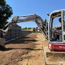 IZDEBSKI - Operator w kabinie mini koparki Takeuchi TB153 wyrównuje teren wzdłuż nowego ogrodzenia panelowego na tle zieleni i błękitnego nieba.