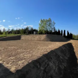 Wyrównany teren pod budowę z widoczną skarpą i przygotowaną drogą dojazdową, drzewa w tle, słoneczny dzień.