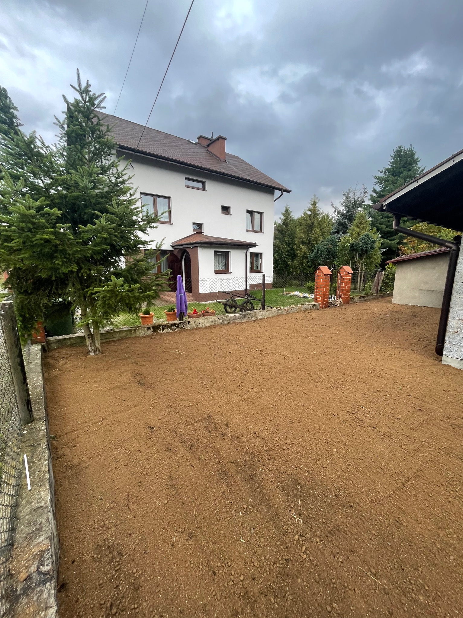 Przygotowany teren pod trawnik przed domem jednorodzinnym, świeżo wyrównana ziemia, widoczne ślady po grabiach, w tle biały dom z brązowym dachem i elementami dekoracyjnymi.