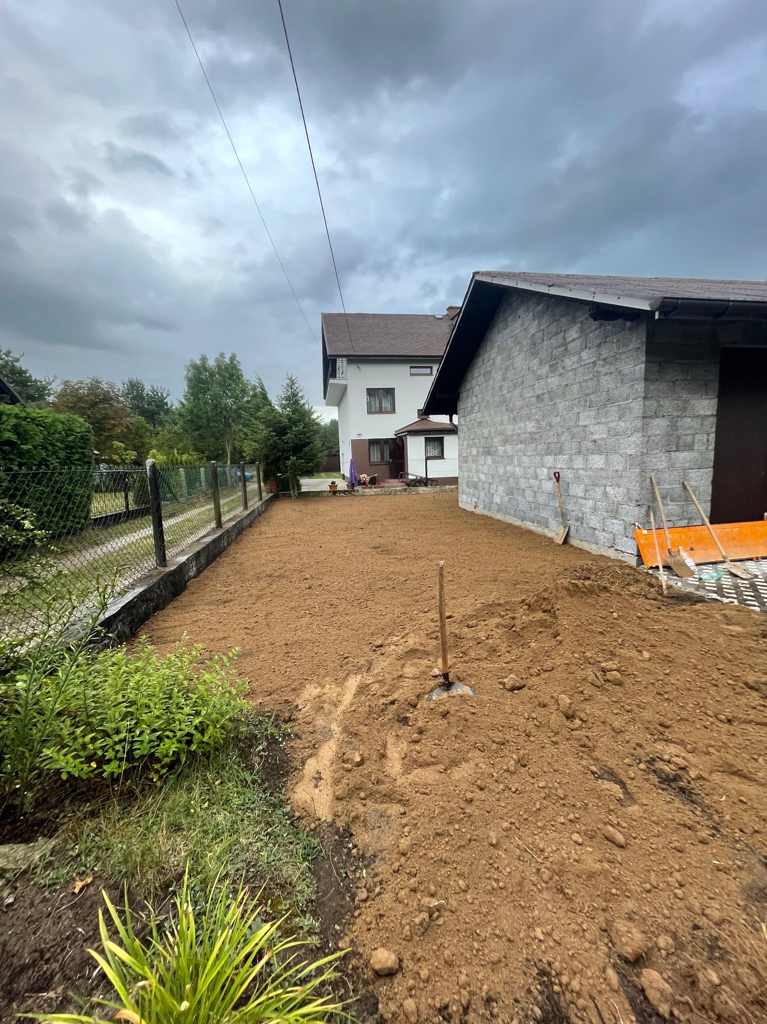 Przygotowany teren pod budowę z widocznymi śladami pracy, narzędziami opartymi o ścianę garażu i pochmurnym niebem w tle.