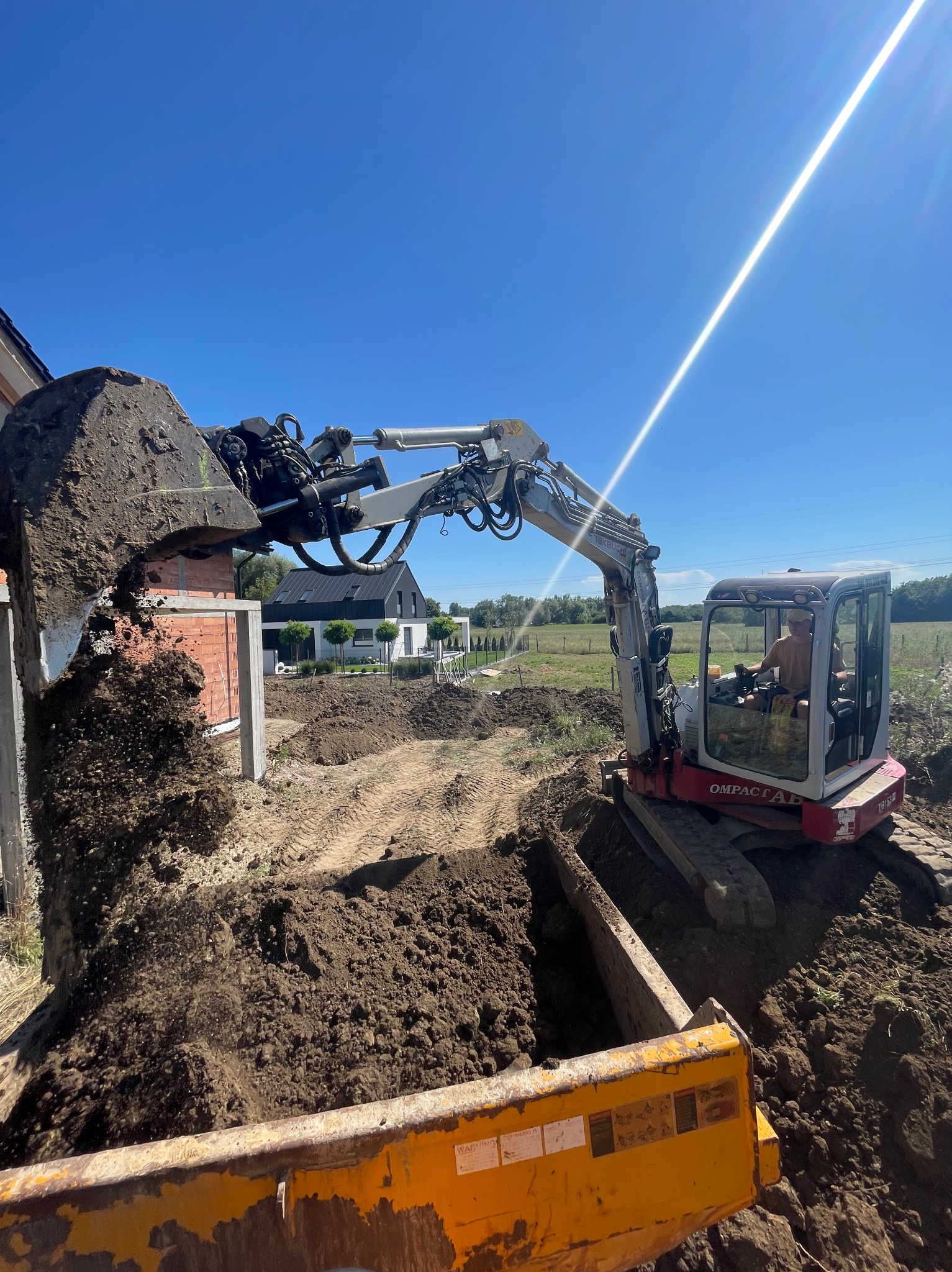 Mała koparka gąsienicowa z operatorem, wysypuje ziemię z wykopu do żółtej przyczepy, w tle budynek w trakcie budowy z czerwonej cegły i dom jednorodzinny.