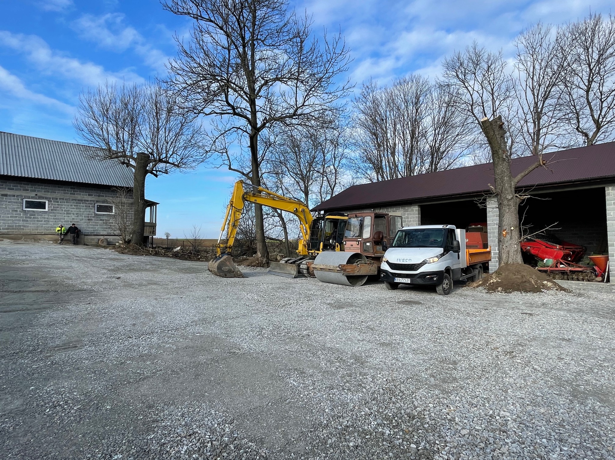 Żółta koparka, walec i biała ciężarówka Iveco na utwardzonym żwirowym placu budowy, w tle budynki gospodarcze i drzewa bez liści, pochmurne niebo.