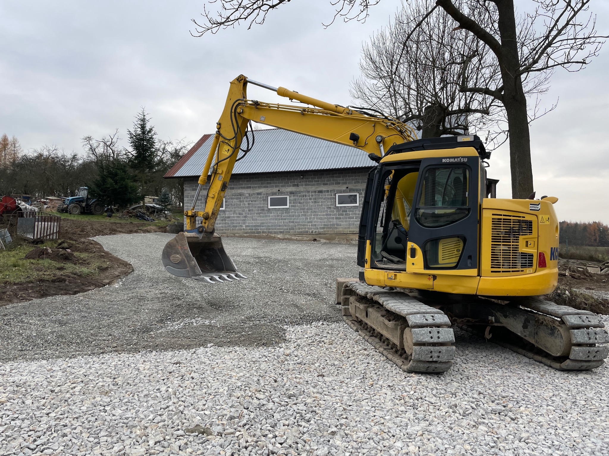 Żółta koparka Komatsu na żwirowej drodze, w tle budynek gospodarczy z szarej cegły i drzewa bez liści, pochmurne niebo.