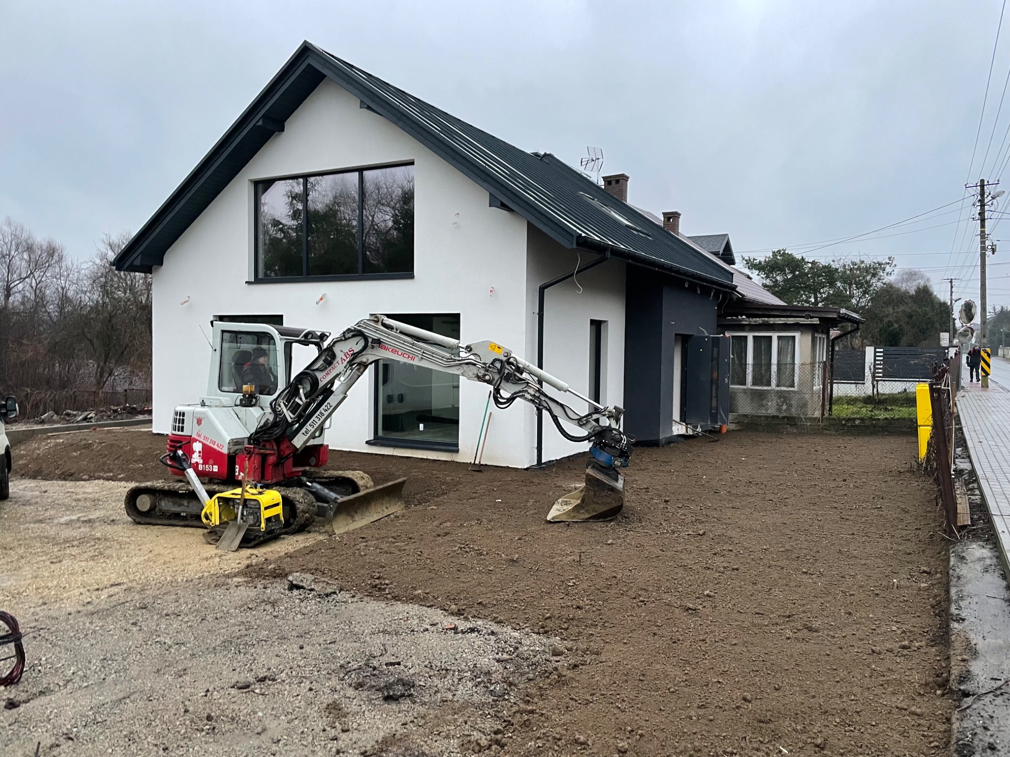 Mini koparka Takeuchi w trakcie prac ziemnych na działce przed nowoczesnym domem z dużym oknem, widoczny fragment utwardzonej drogi i świeżo rozplantowana ziemia.
