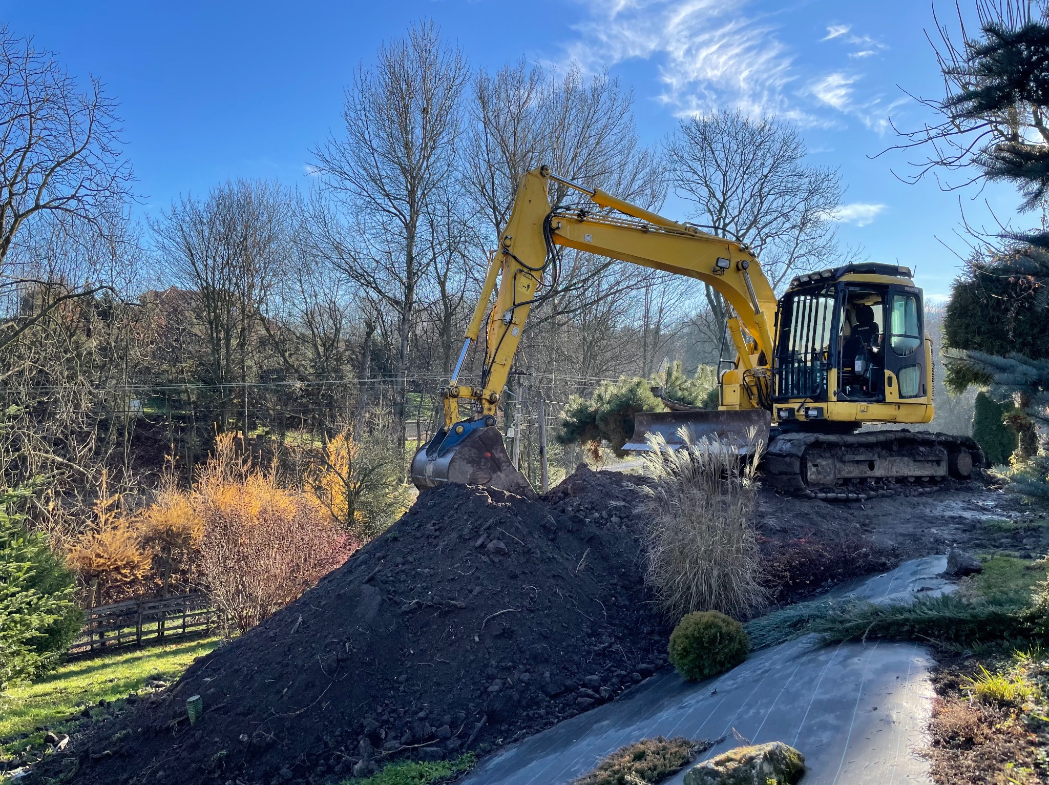 Żółta koparka nabiera ziemię na usypany kopiec na tle drzew bez liści i błękitnego nieba.