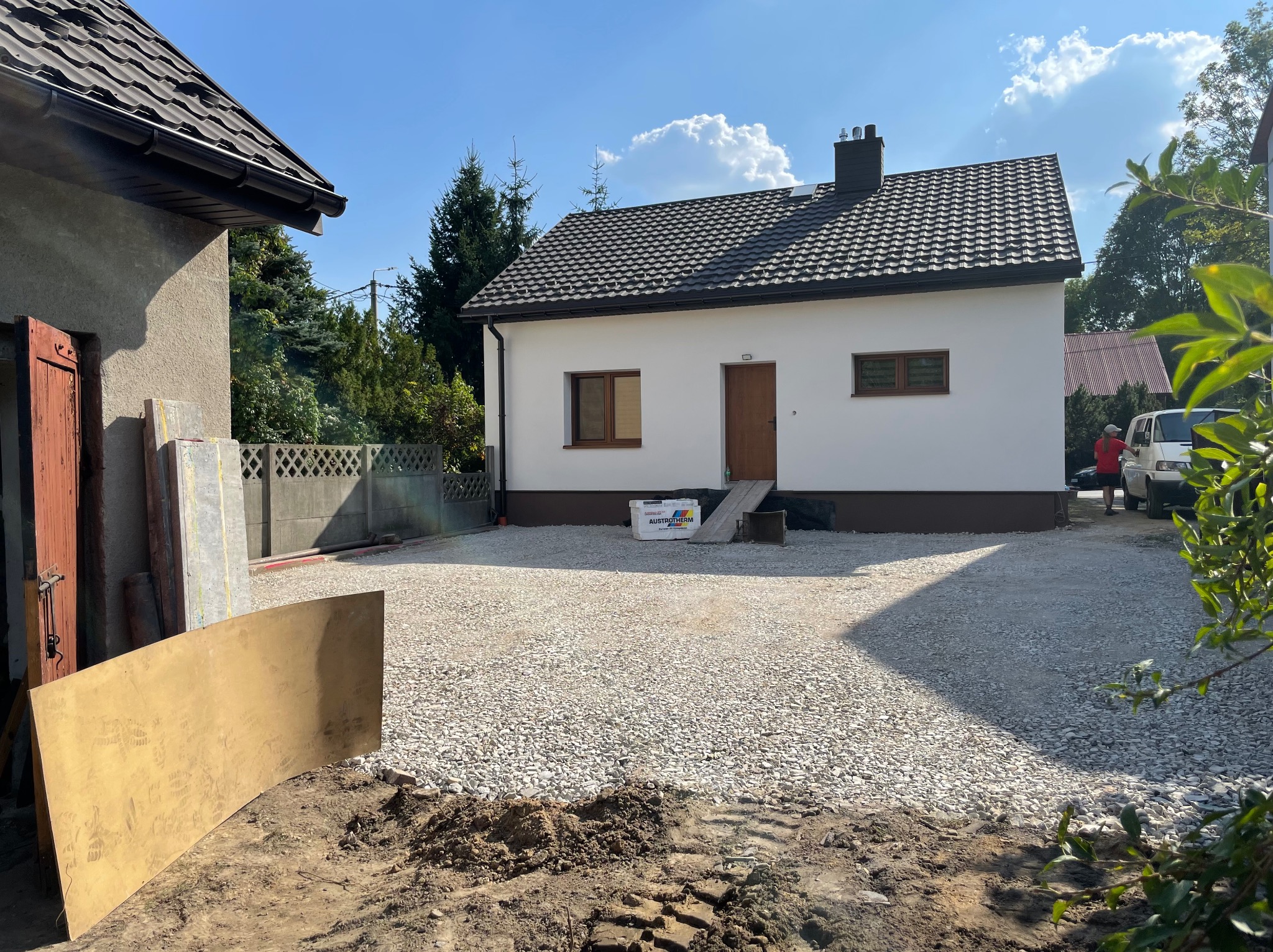Podwórko z nowo wysypanym żwirem przed białym domem z brązowymi oknami i drzwiami, widoczne ślady opon i deska oparta o ścianę budynku gospodarczego.