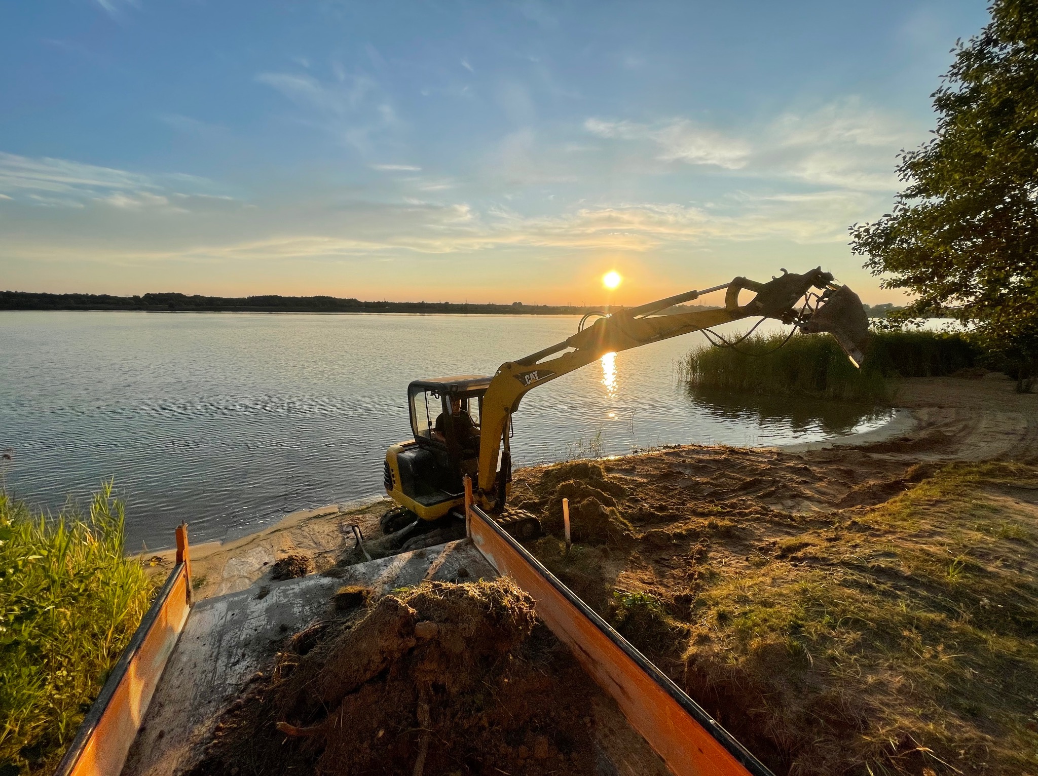 Żółta minikoparka CAT nabiera ziemię przy brzegu jeziora o zachodzie słońca; widoczna część przyczepy i drzewa w tle.