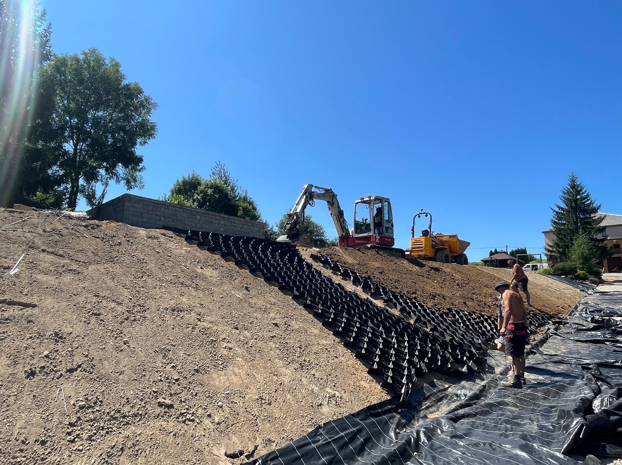 Skarpa wzmocniona geokratą i zabezpieczona geowłókniną, w tle minikoparka i wywrotka, dwóch mężczyzn przy pracy.