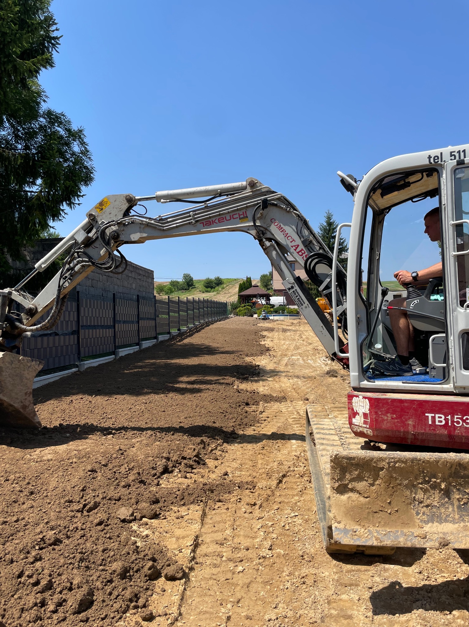 Operator w kabinie mini koparki Takeuchi TB153 wyrównuje teren wzdłuż nowego ogrodzenia panelowego na tle zieleni i błękitnego nieba.