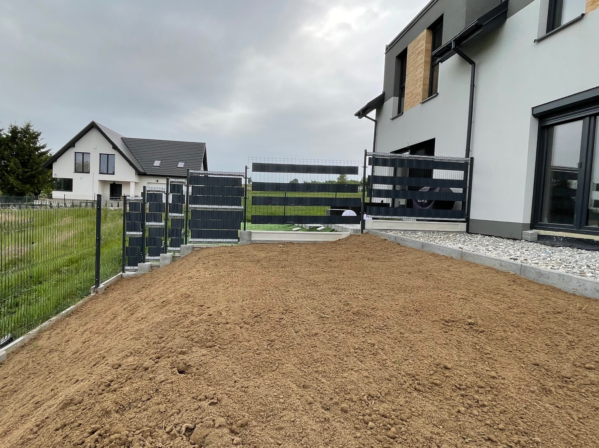 Przygotowany teren pod trawnik z nawiezioną ziemią, obrzeża z kostki, nowoczesne domy w tle, pochmurne niebo.