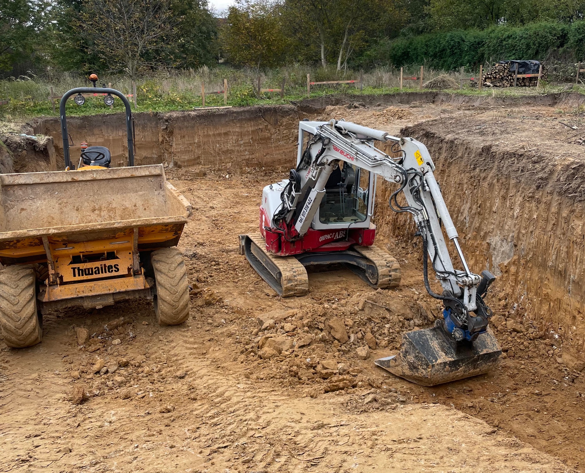 Żółta wywrotka Thwaites i biało-czerwona koparka Takeuchi w wykopie pod fundamenty, widoczne ślady pracy w ziemi.