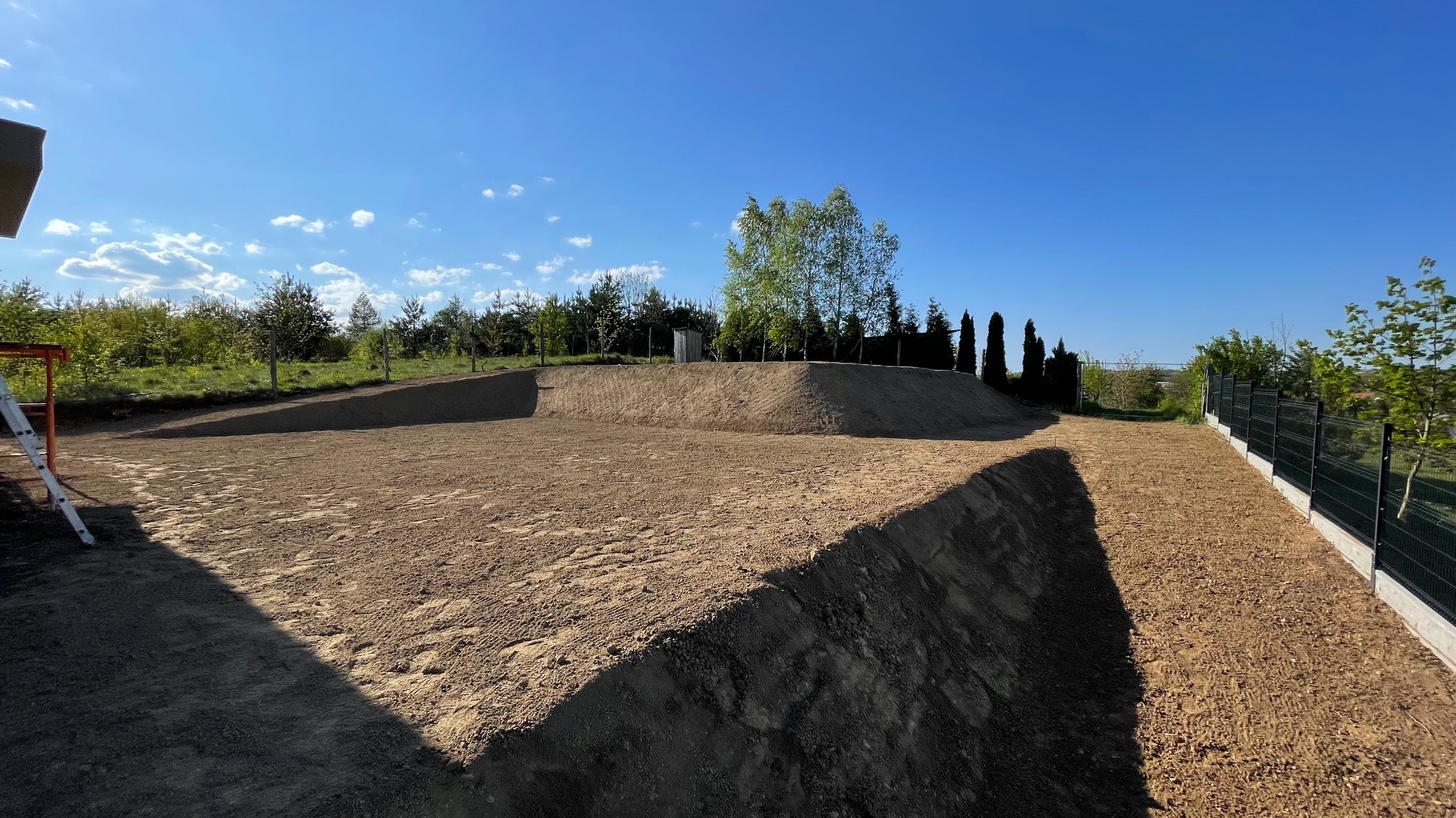 Wyrównany teren pod budowę z widoczną skarpą i przygotowaną drogą dojazdową, drzewa w tle, słoneczny dzień.