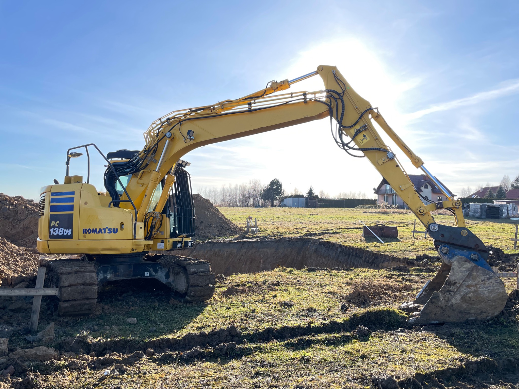 Żółta koparka Komatsu PC138US w trakcie wykonywania wykopu w ziemi na tle błękitnego nieba i słońca.
