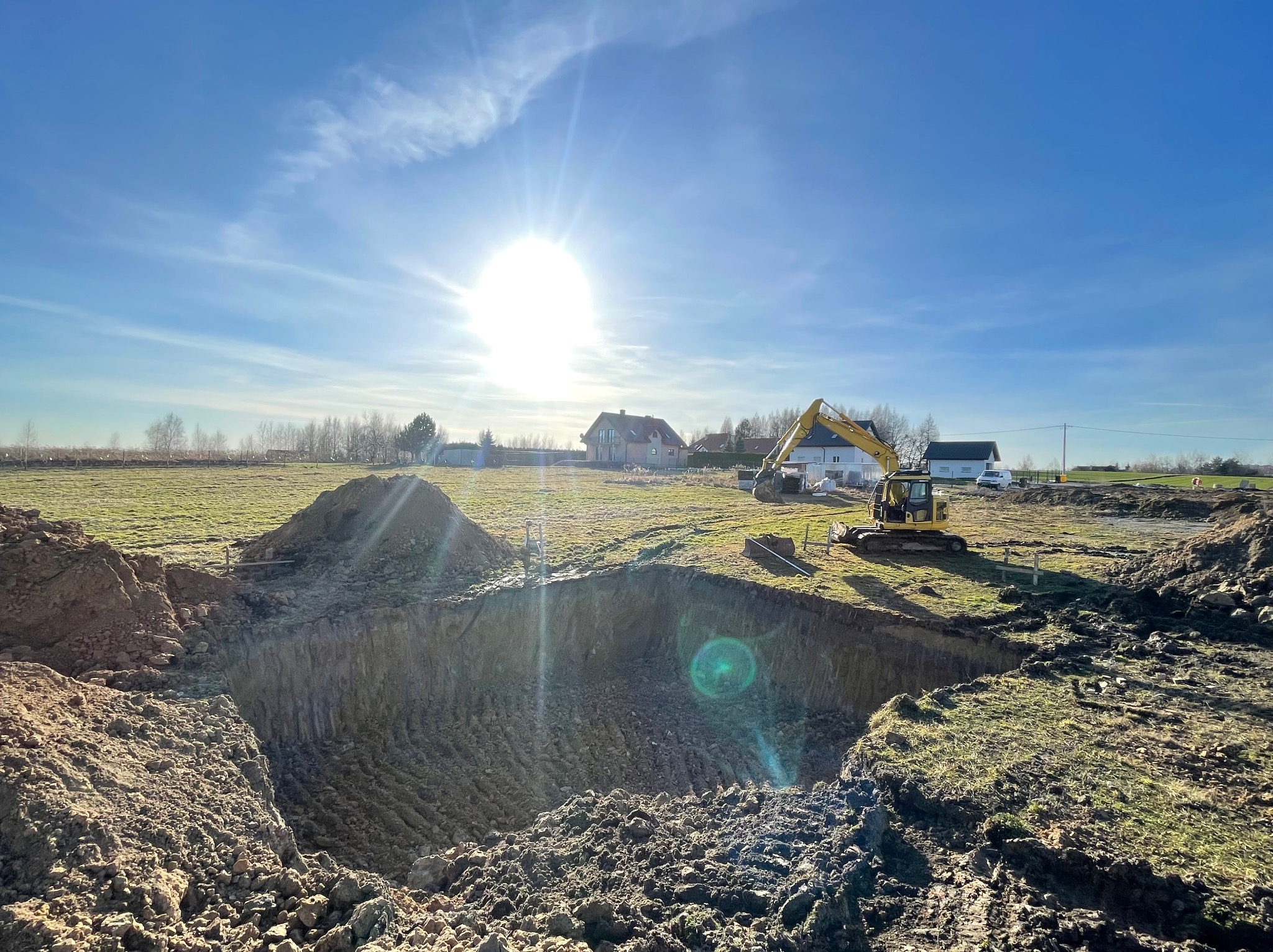 Głęboki wykop w ziemi na słonecznym polu, z widoczną żółtą koparką i zabudowaniami w tle.