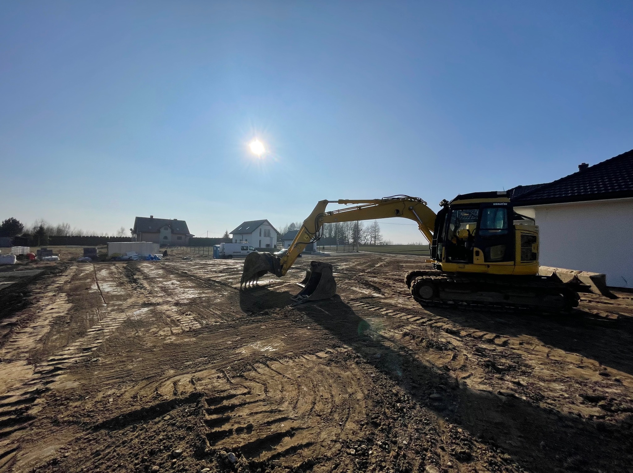 Żółta koparka Komatsu na wyrównanym, przygotowanym pod budowę terenie z widocznymi śladami opon, w tle domy jednorodzinne i błękitne niebo z silnym słońcem.