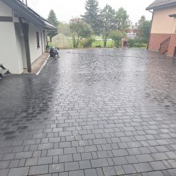 Firma Usługowa 3K - Ułożona, mokra, grafitowa kostka brukowa na posesji, widoczny pracownik w trakcie układania przy budynku z zewnętrznymi schodami, w tle drugi budynek i zieleń.