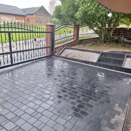 Firma Usługowa 3K - Ułożona, mokra, grafitowa kostka brukowa przed domem z ozdobną, kutą bramą w tle, widoczny drenaż liniowy.