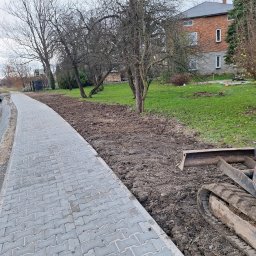 Firma Usługowa 3K - Świeżo ułożony chodnik z szarej kostki brukowej sąsiaduje z przygotowanym terenem pod nasadzenia, gdzie widać ślady pracy koparki; w tle budynek mieszkalny i drzewa.