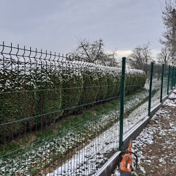 Firma Usługowa 3K - Zielone, metalowe ogrodzenie panelowe z podmurówką biegnące wzdłuż zimozielonego żywopłotu pokrytego śniegiem, z pomarańczową pilarką spalinową leżącą u podstawy słupka.