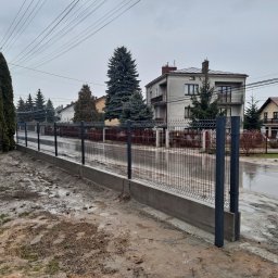 Firma Usługowa 3K - Nowo zamontowane, panelowe ogrodzenie z podmurówką betonową w kolorze grafitowym na tle szarego, pochmurnego nieba i zabudowy jednorodzinnej po deszczu.