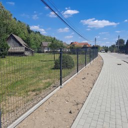 Firma Usługowa 3K - Nowo zamontowane, grafitowe ogrodzenie panelowe z podmurówką betonową, oddzielające posesję od chodnika wyłożonego szarą kostką brukową. W tle wiejska zabudowa i błękitne niebo.