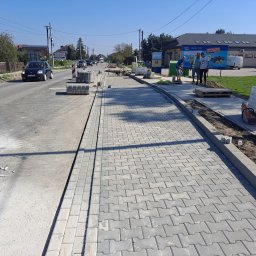 Firma Usługowa 3K - Nowy chodnik z kostki brukowej w Jodłówce, w trakcie układania. Widoczni pracownicy, maszyna do cięcia kostki, palety z kostką i ruch uliczny w tle.