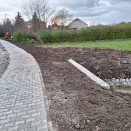 Firma Usługowa 3K - Świeżo ułożona, szara kostka brukowa wzdłuż drogi z widocznym wykopem pod rzekę, z pracującym w tle małym traktorem i robotnikiem w żółtej kamizelce, przy pochmurnym niebie.