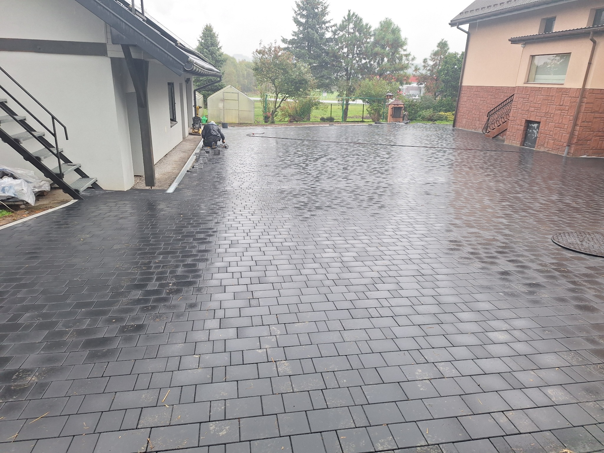 Ułożona, mokra, grafitowa kostka brukowa na posesji, widoczny pracownik w trakcie układania przy budynku z zewnętrznymi schodami, w tle drugi budynek i zieleń.