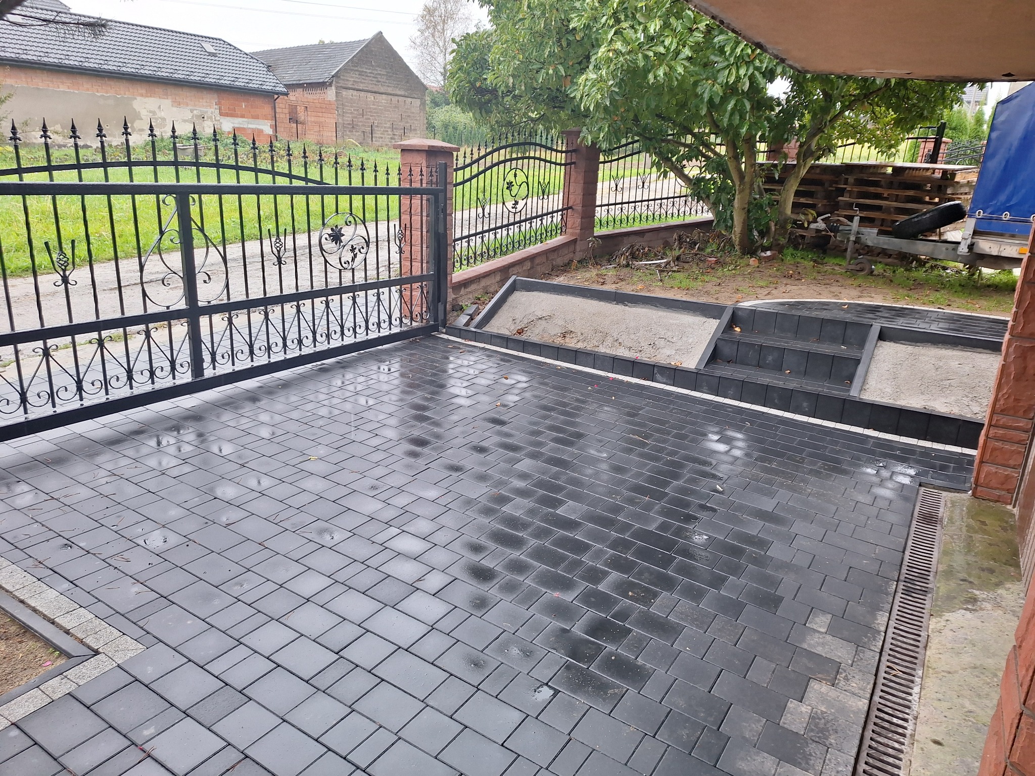 Ułożona, mokra, grafitowa kostka brukowa przed domem z ozdobną, kutą bramą w tle, widoczny drenaż liniowy.