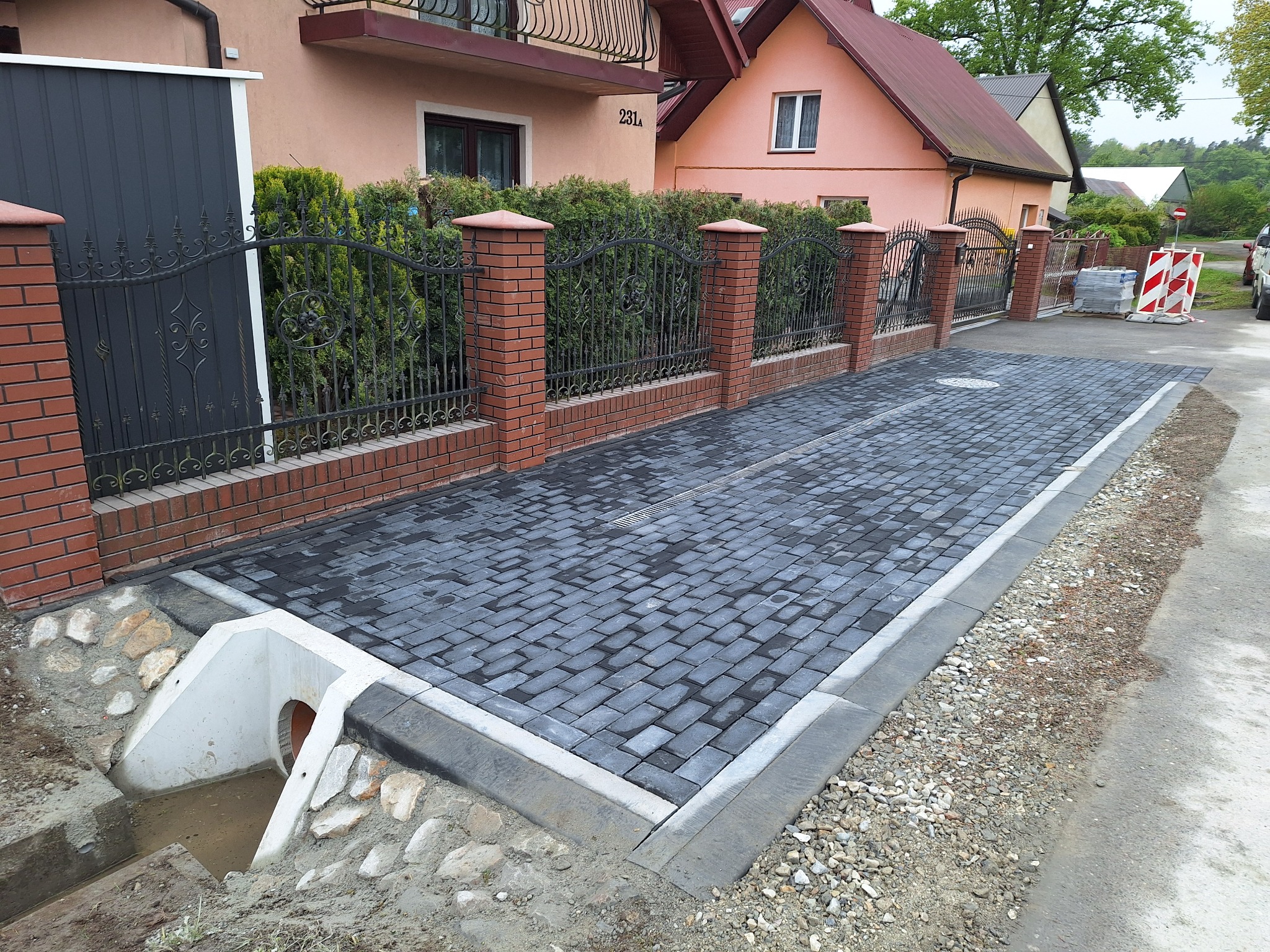 Nowo ułożona, szara kostka brukowa przed posesją z ceglanym ogrodzeniem i ozdobną bramą, widoczny betonowy odpływ wody deszczowej z rurą. Dom z numerem 231a w tle.