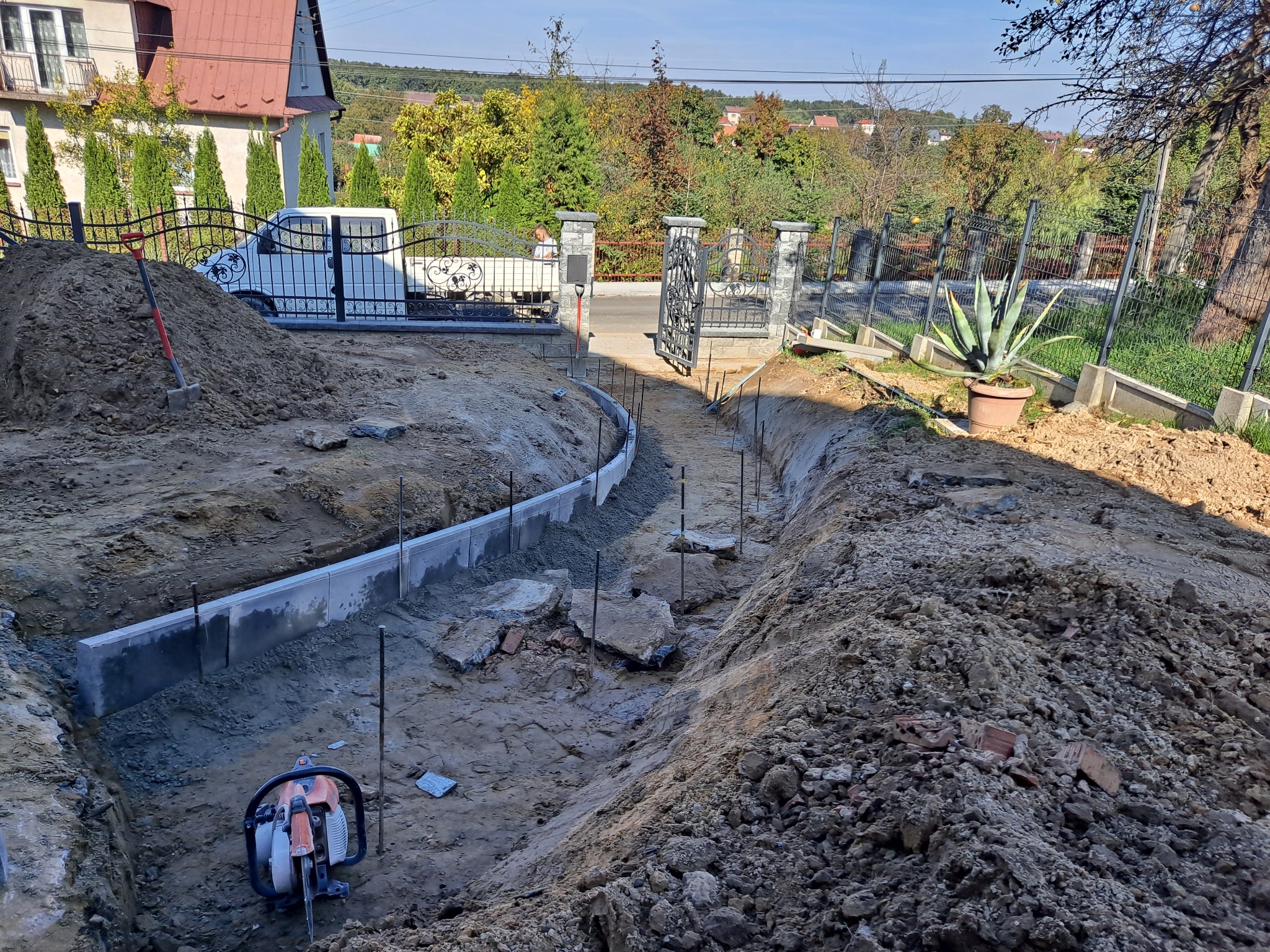Przygotowanie podłoża pod ścieżkę z betonowymi obrzeżami na posesji, widoczny wykop, paliki stabilizujące, piła do cięcia betonu, brama wjazdowa i zaparkowany samochód dostawczy w tle.
