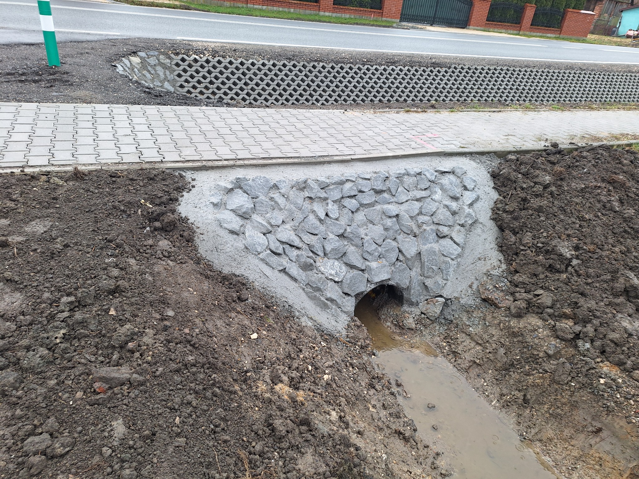 Przepust drogowy z rurą, wzmocniony kamieniami i betonem, odprowadzający wodę spod chodnika i drogi. Widoczna ziemia, chodnik z kostki i fragment drogi.