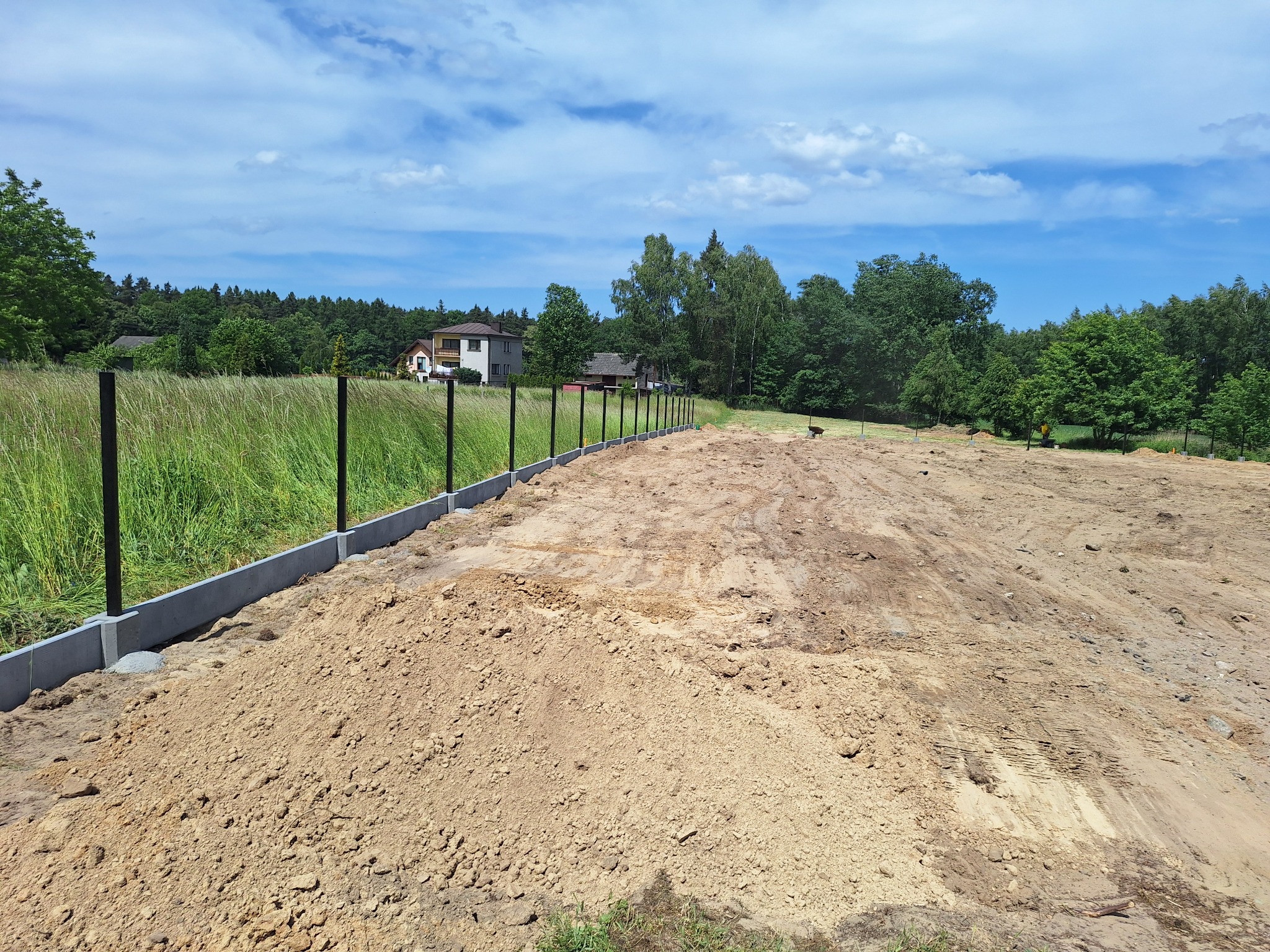 Przygotowany teren pod budowę, widoczne słupki i betonowe obrzeża, w tle domy i zieleń.