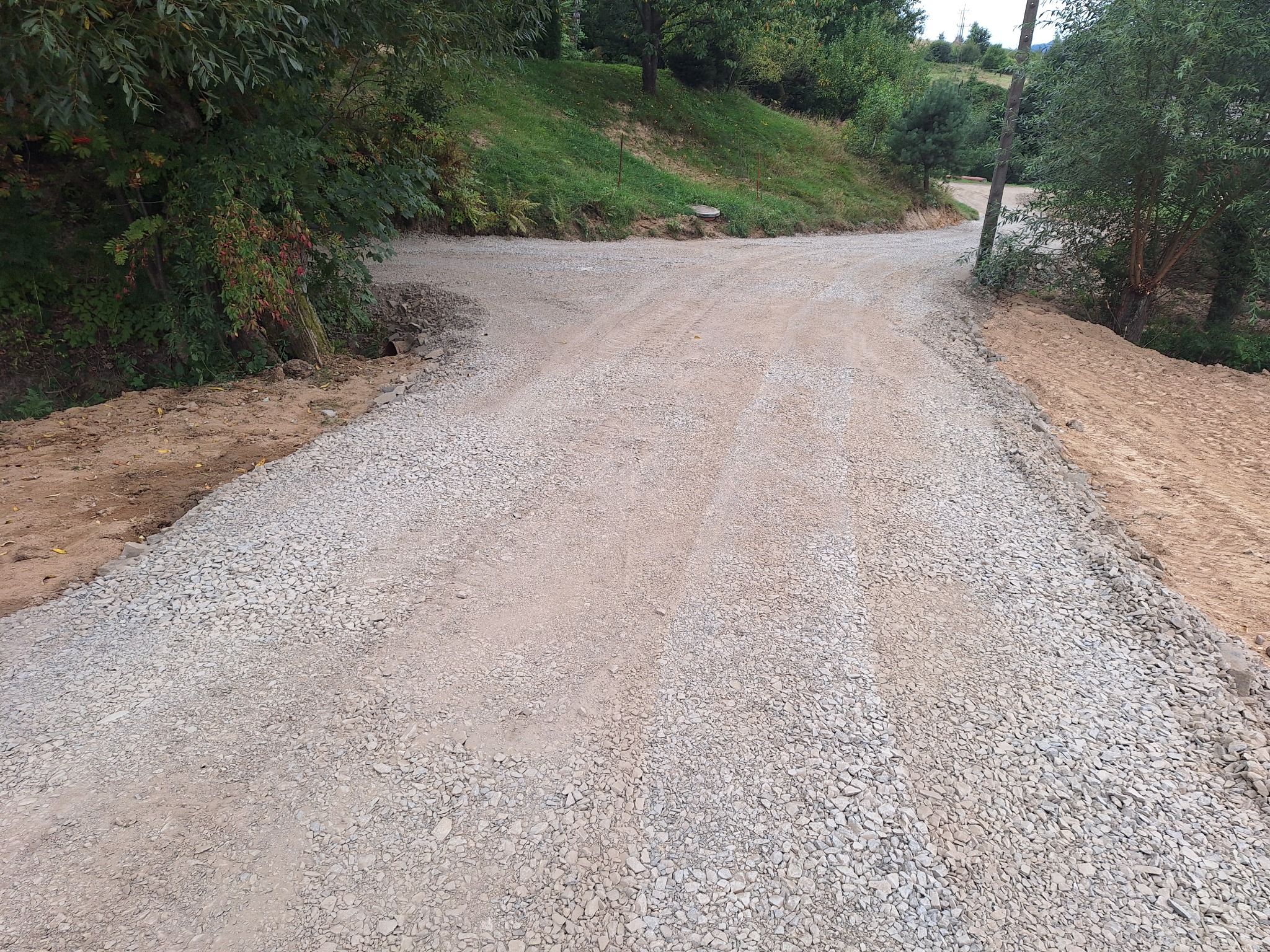 Szeroki kadr na świeżo utwardzoną, szutrową drogę gruntową na rozwidleniu, otoczoną zielenią drzew i krzewów, z widocznymi śladami opon.
