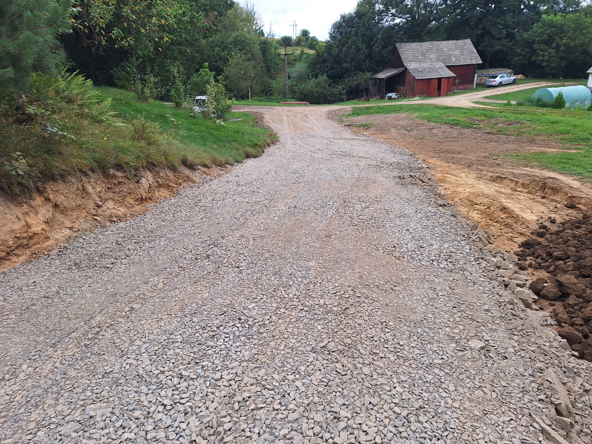Utwardzona droga gruntowa z szarym kruszywem, widoczne ślady opon, brązowa ziemia po bokach, w tle budynek gospodarczy i zieleń.