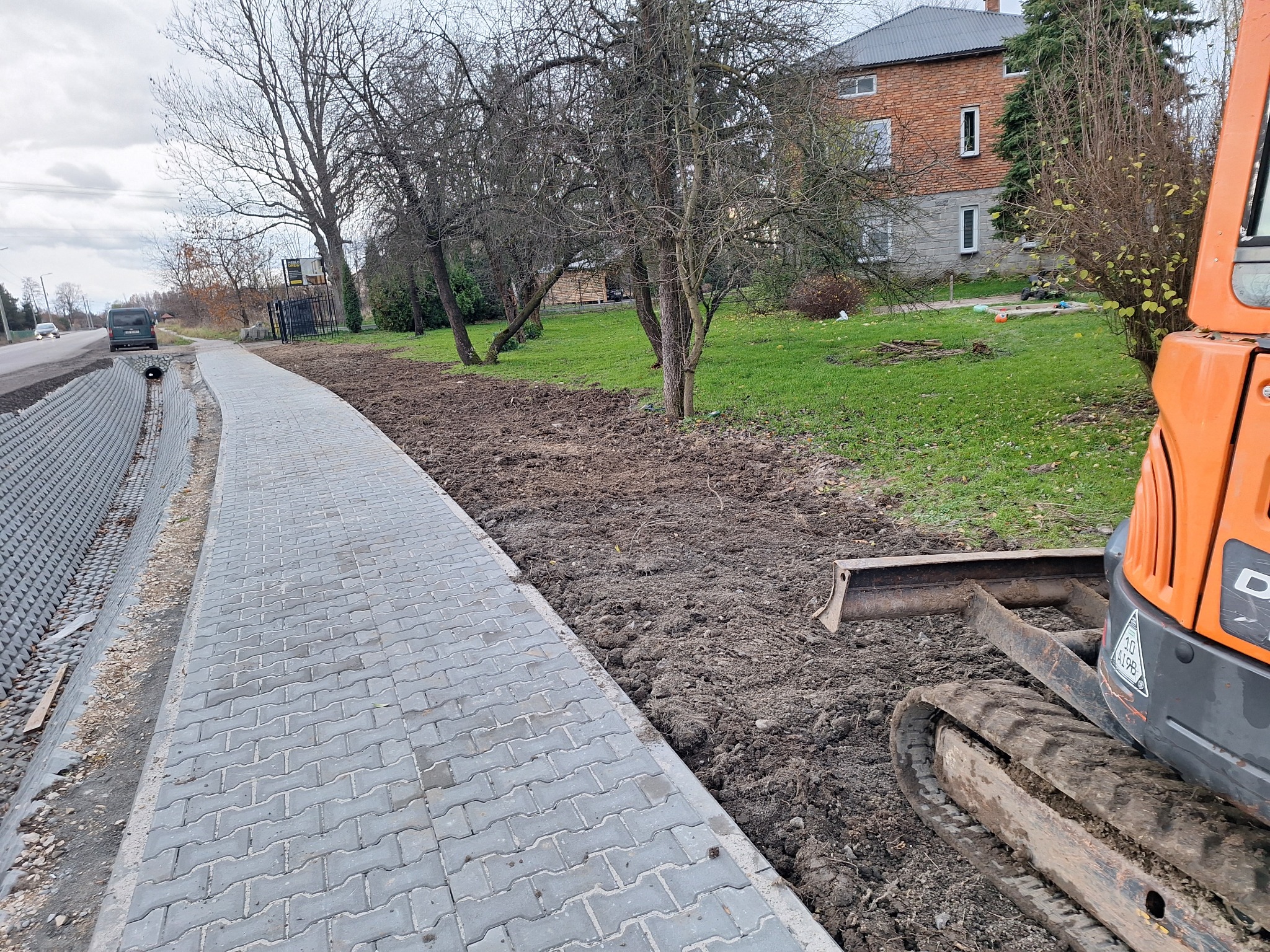 Świeżo ułożony chodnik z szarej kostki brukowej sąsiaduje z przygotowanym terenem pod nasadzenia, gdzie widać ślady pracy koparki; w tle budynek mieszkalny i drzewa.