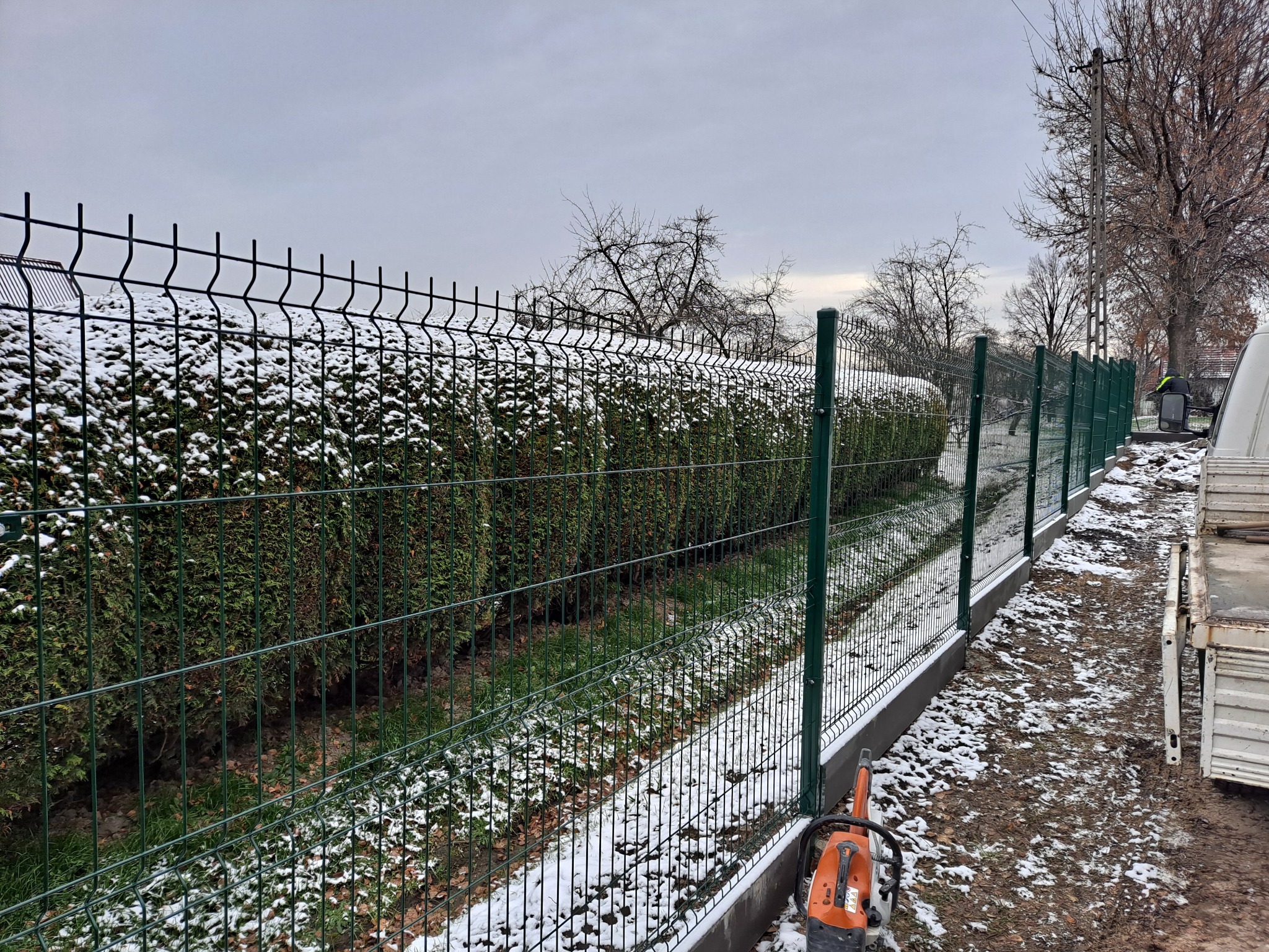 Zielone, metalowe ogrodzenie panelowe z podmurówką biegnące wzdłuż zimozielonego żywopłotu pokrytego śniegiem, z pomarańczową pilarką spalinową leżącą u podstawy słupka.