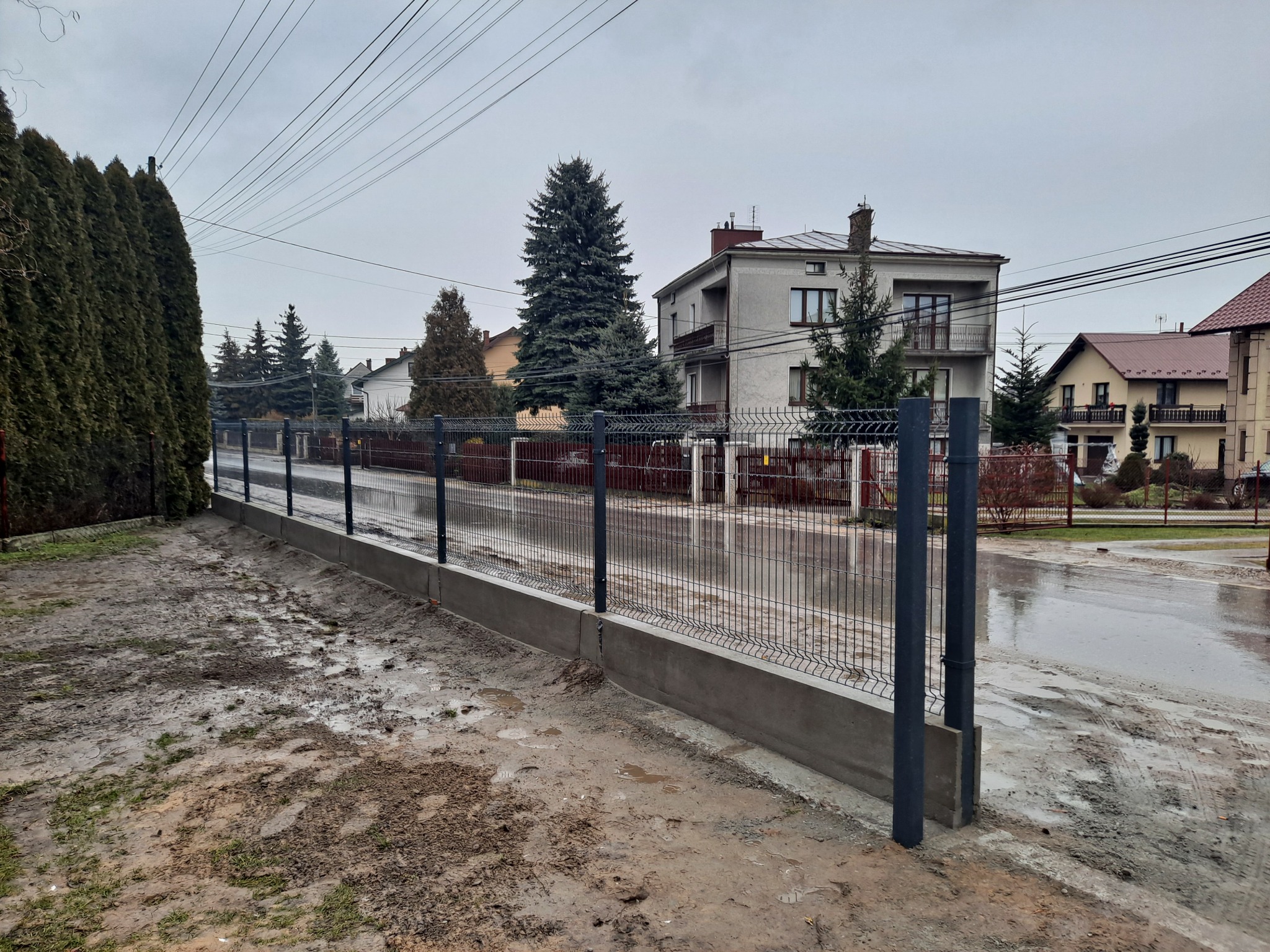 Nowo zamontowane, panelowe ogrodzenie z podmurówką betonową w kolorze grafitowym na tle szarego, pochmurnego nieba i zabudowy jednorodzinnej po deszczu.
